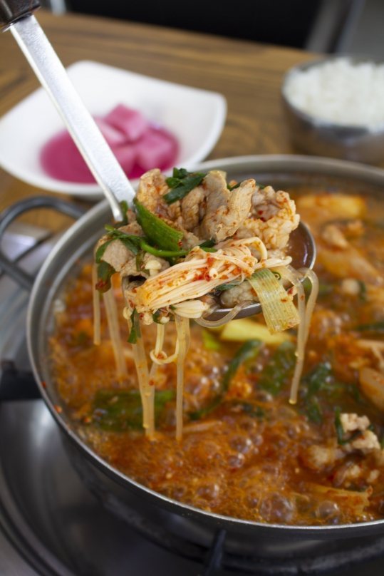 버너 위에서 보글보글 끓는 냄비에서 국자로 떠 올린 와촌돼지찌개와 공기밥, 무절임